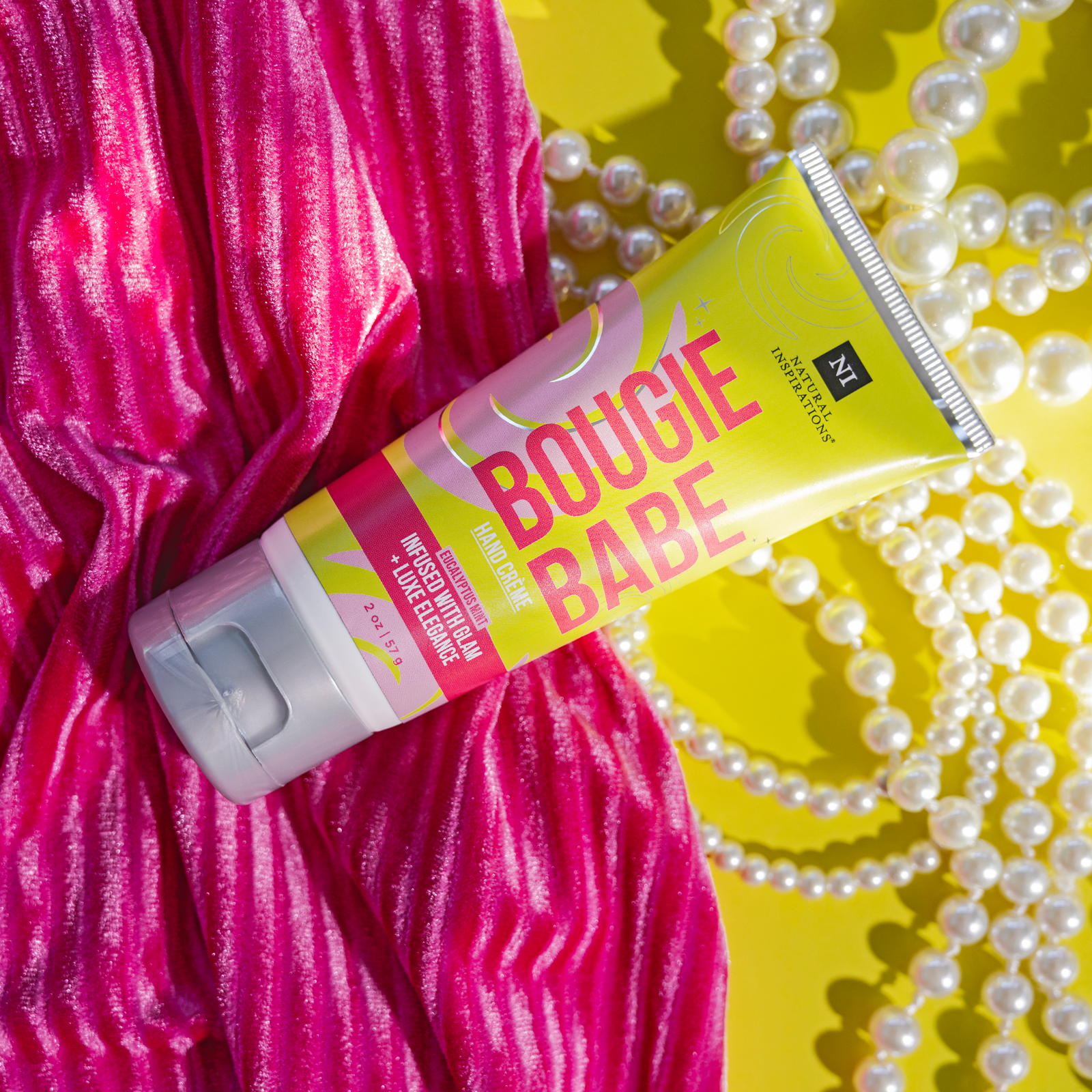 close up of bougie babe mini hand cream on pink fabric, a green background and pearls scattered throughout
